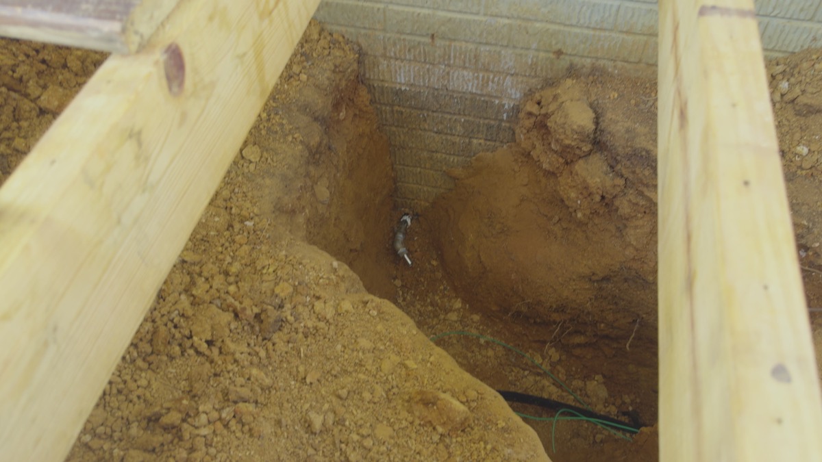 Excavated access opening with exposed pipe beside a brick foundation
