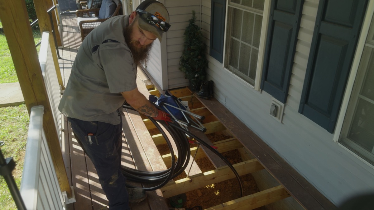 FOX crew member handling coiled black pipe beside an opened porch floor